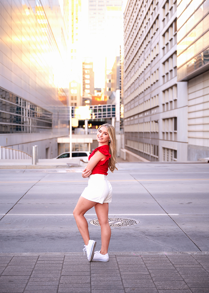 A senior girl in a red top and white shorts stands confidently on an empty city street at sunset, with tall buildings reflecting a warm glow.