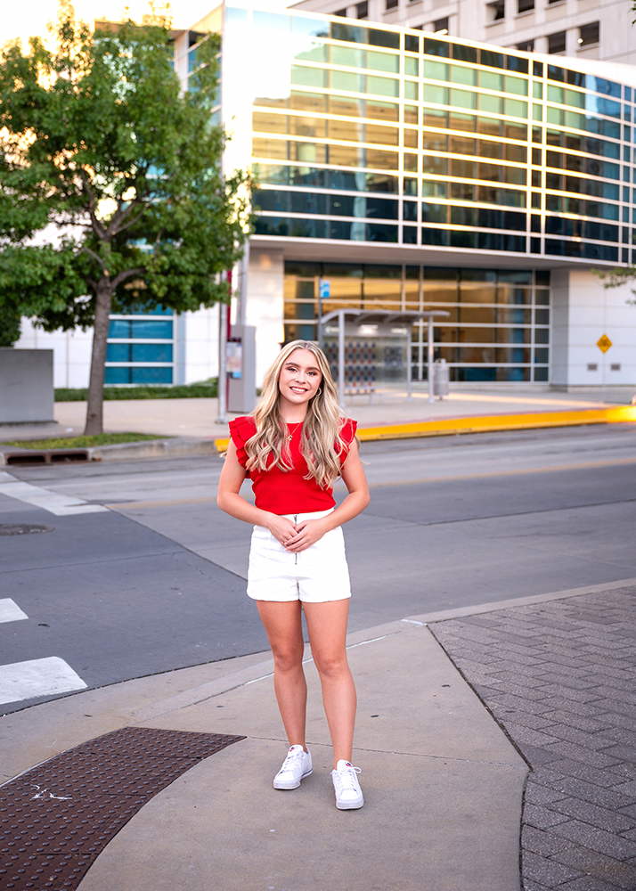 Senior photos of Grace wearing a red top and white shorts while standing on an urban street. Glass building and tree in the background on a sunny day.
