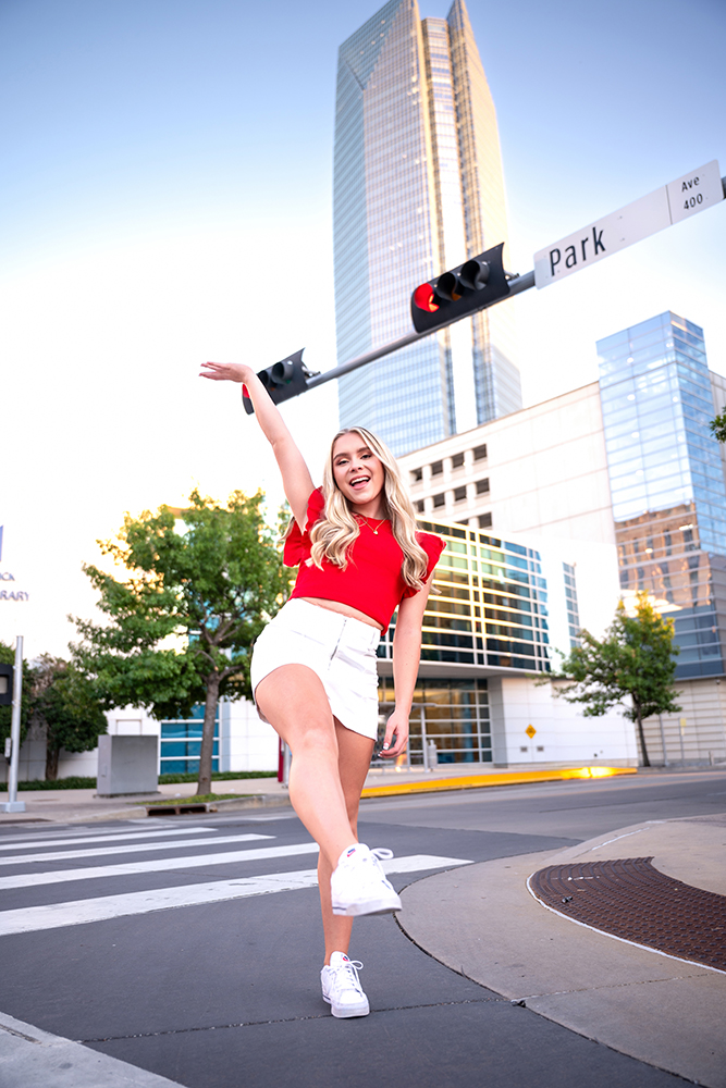 High school senior in a red shirt and white skirt joyfully poses on a city crosswalk. A tall, mirrored skyscraper and traffic lights are visible in the background.