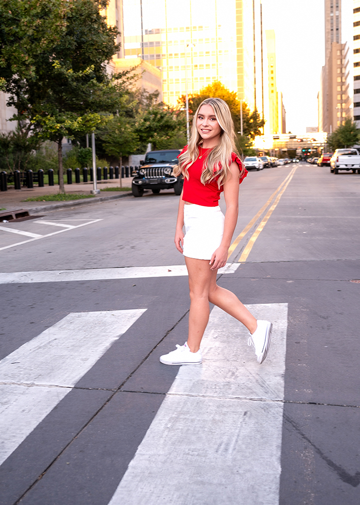 High school senior with long blonde hair, wearing a red top and white skirt, smiles while walking across a city street crosswalk at sunset. Urban and cheerful.