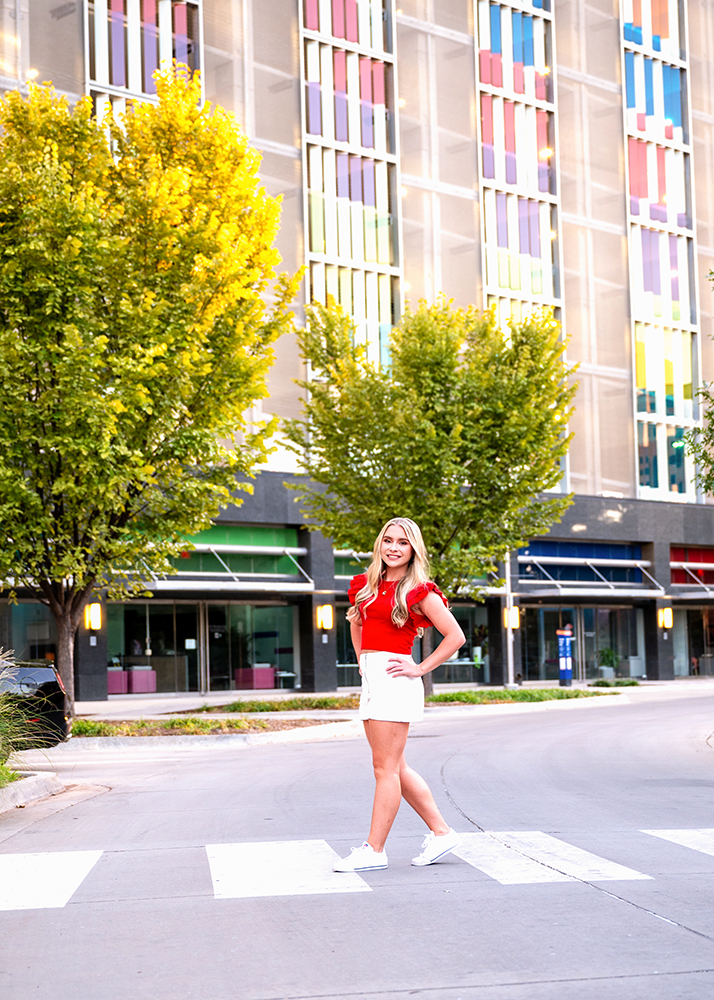 Senior photos of a girl in a red top and white skirt stands confidently on a crosswalk, with vibrant, colorful windows and lush green trees in the background.