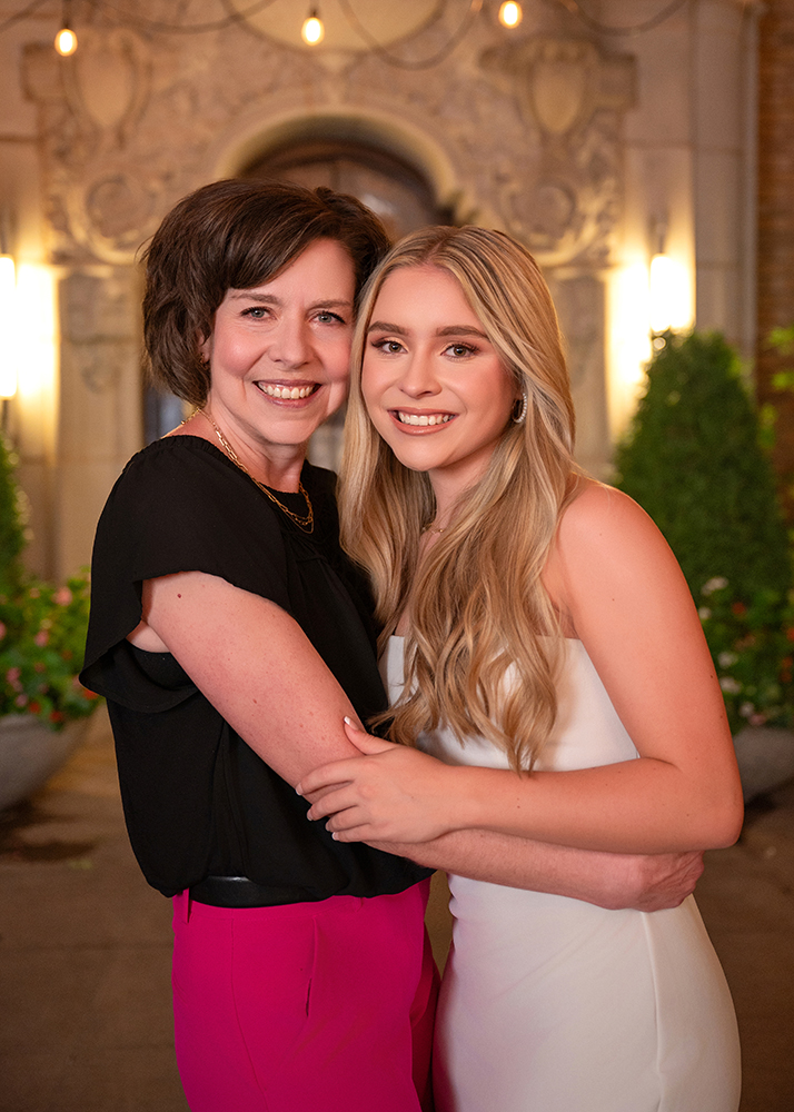 A smiling woman in a black top and pink pants embraces a young woman in a white dress. They're in a warmly lit, elegant courtyard setting.