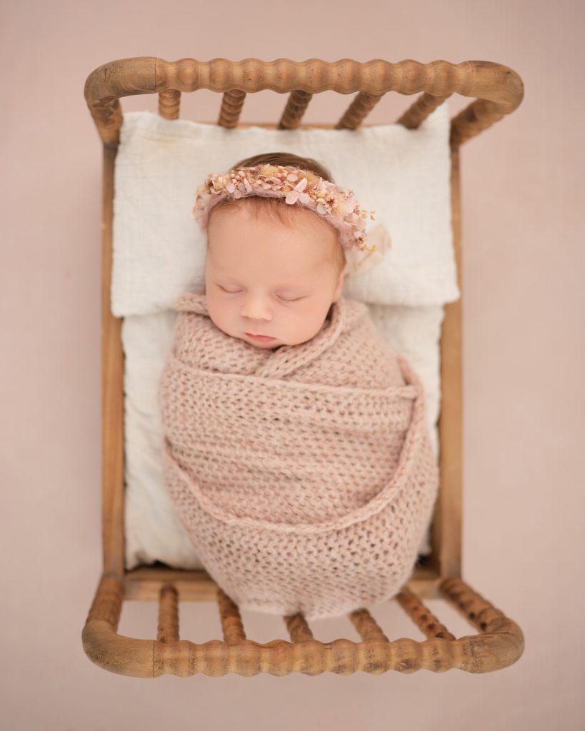 A sleeping baby wrapped in a soft pink knit blanket rests on a tiny wooden bed