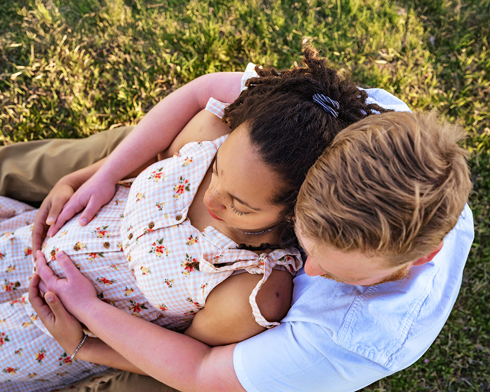 A couple sits on grass, lovingly embracing. The man cradles the woman's pregnant belly as she rests against him, wearing a floral dress.
