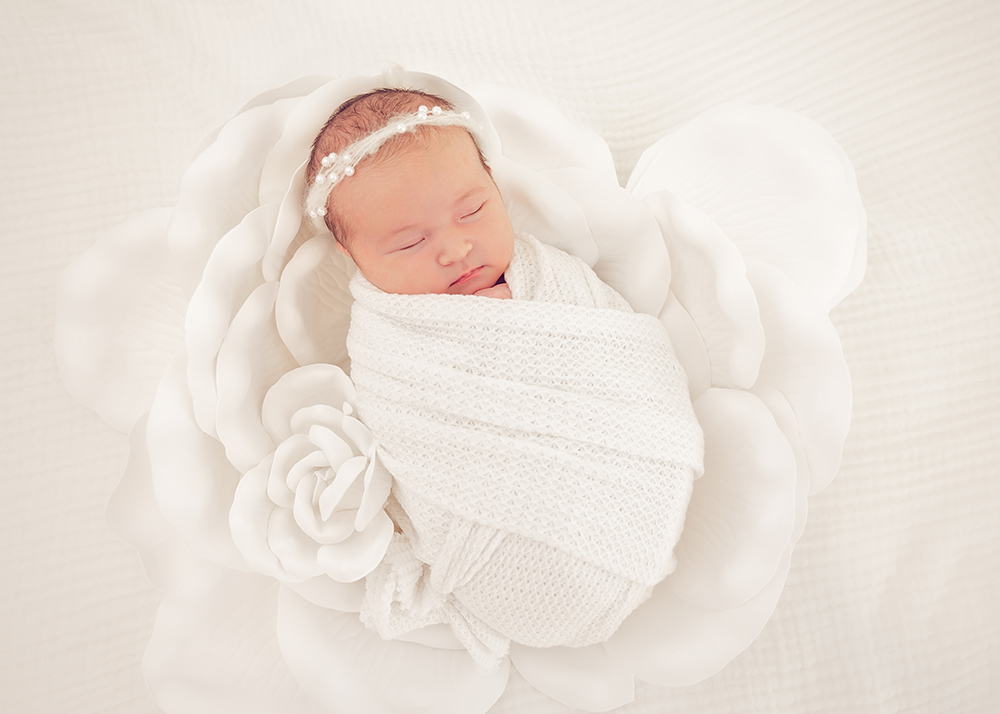 A newborn baby peacefully sleeps wrapped in a textured, white blanket with a delicate pearl headband, nestled atop oversized white flower.