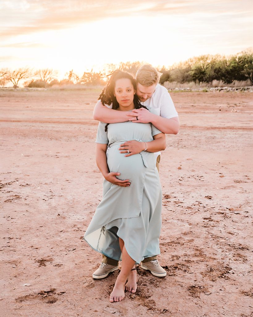 A pregnant woman in a light blue dress stands barefoot on sandy ground, embraced from behind by a partner. The golden sunset creates a warm, peaceful glow.