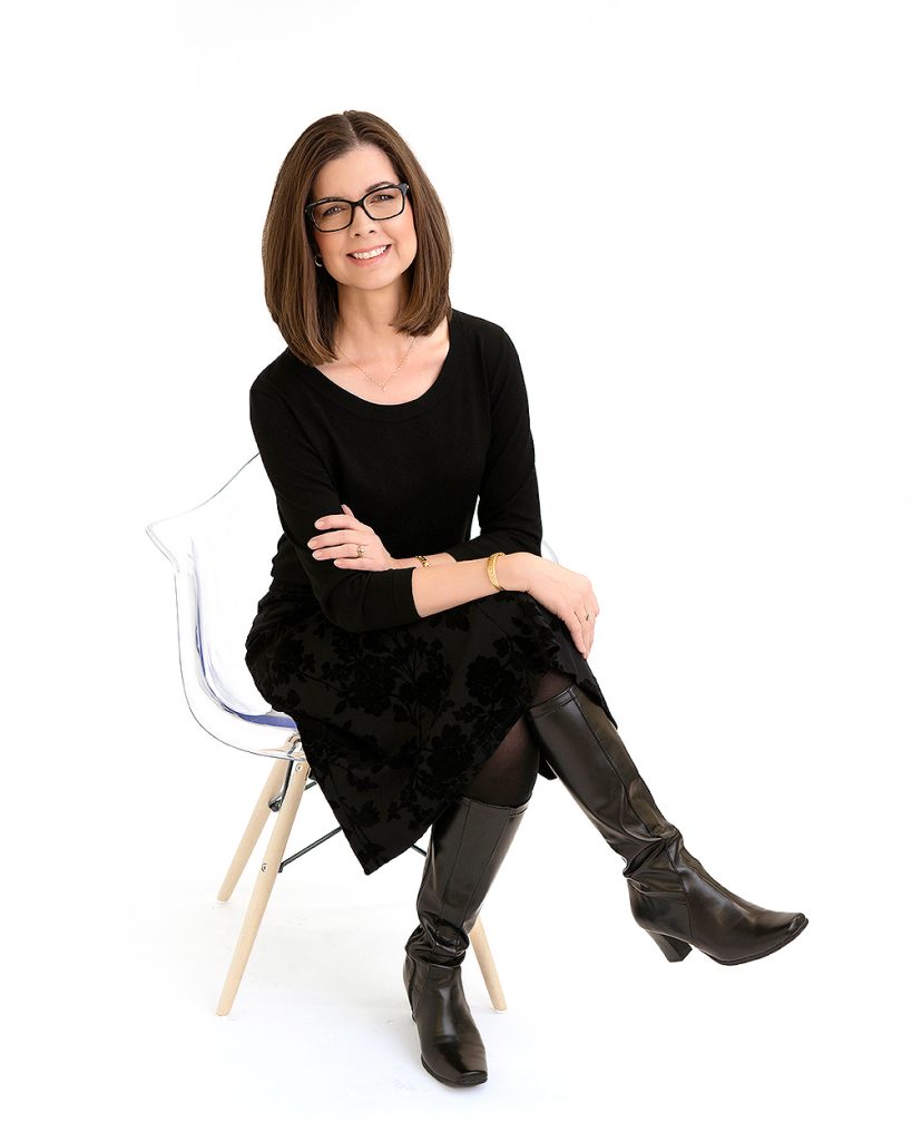A woman with glasses, wearing a black dress and knee-high boots, sits confidently on a clear chair against a white background