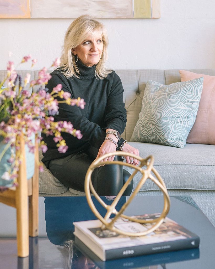 A woman sits on a sofa with pastel cushions, smiling thoughtfully