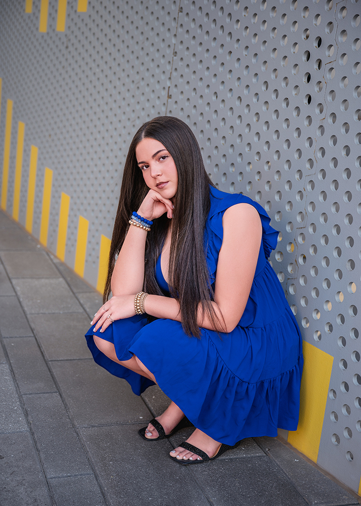 A senior girl in a blue dress crouches beside a perforated metal wall with yellow stripes. She rests her chin on her hand, giving a confident, thoughtful look.