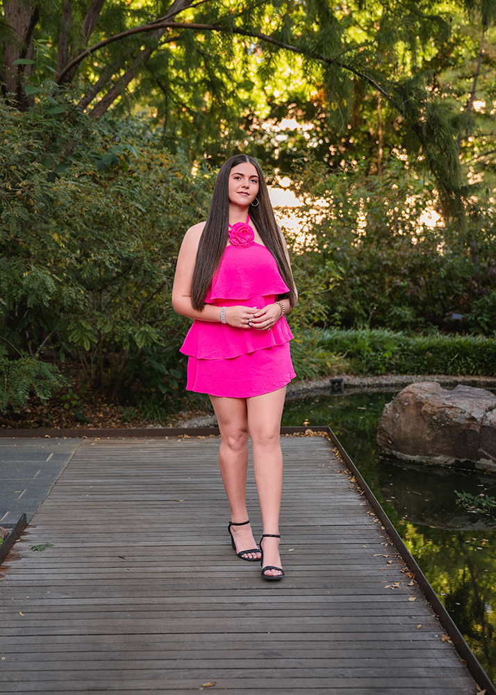 A high school senior in a bright pink dress stands on a wooden walkway surrounded by greenery. She appears calm and confident in a serene outdoor setting.