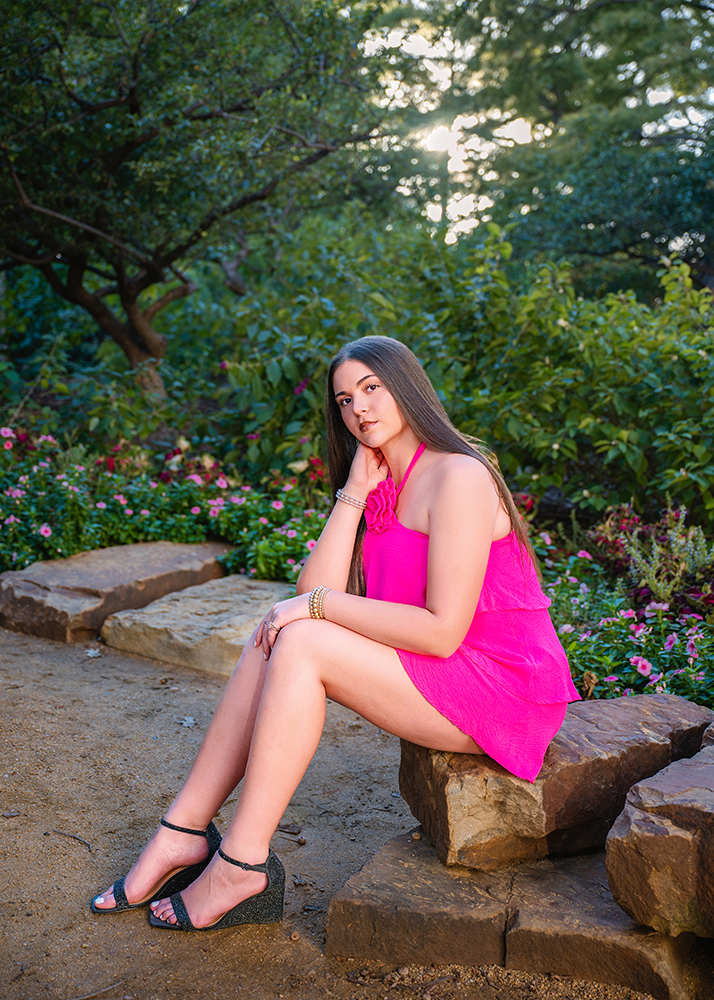 A senior girl in a vibrant pink dress sits on a stone bench in a lush garden, surrounded by colorful flowers. The sun peeks through trees, creating a serene atmosphere.