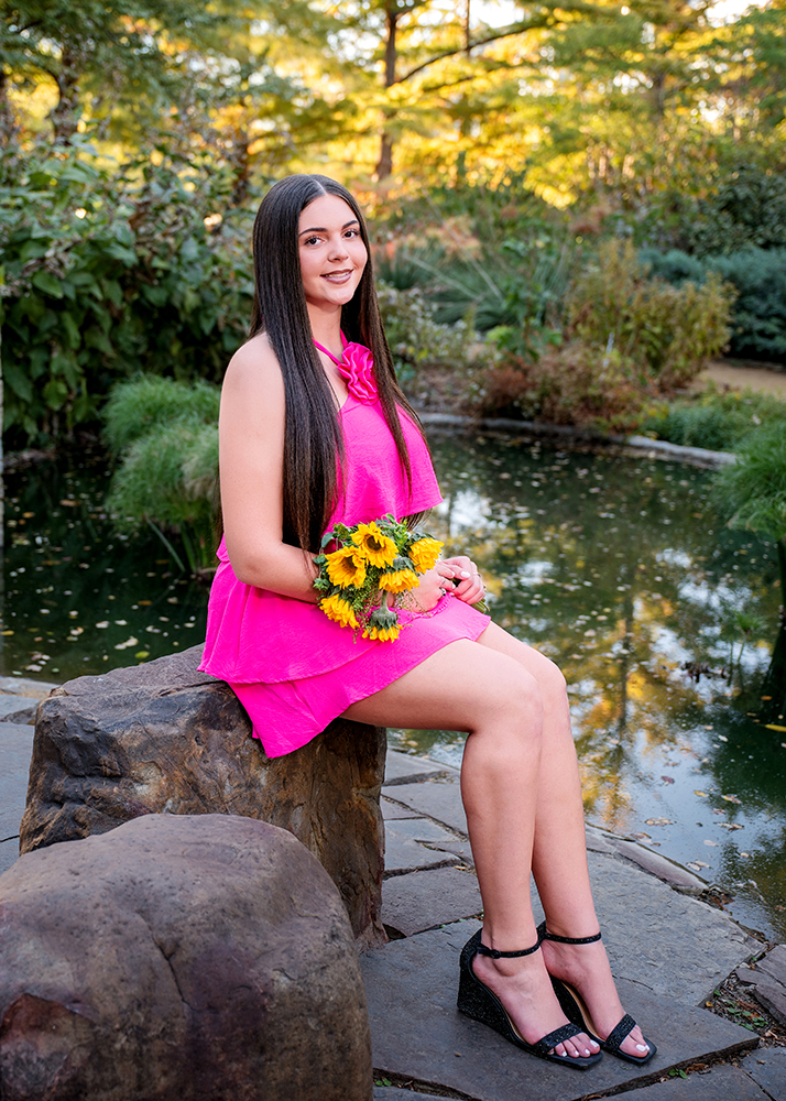 A senior girl in a vibrant pink dress and black heels sits on a rock by a serene pond, holding sunflowers. The scene is peaceful, with lush greenery.