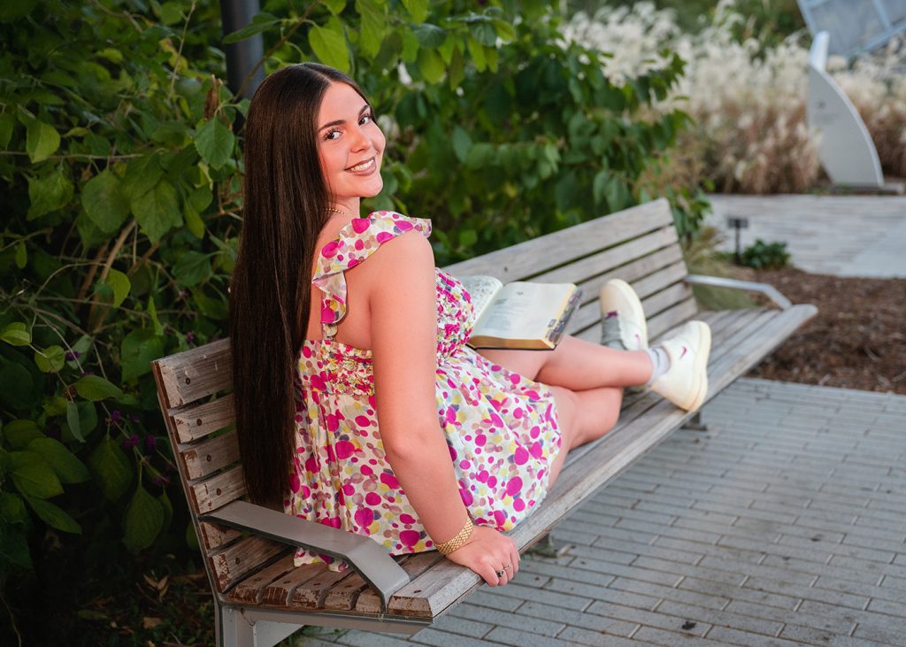 A senior girl sitting on a bench in a garden, wearing a colorful floral dress, holding an open book, and smiling over her shoulder. The scene is serene and joyful.