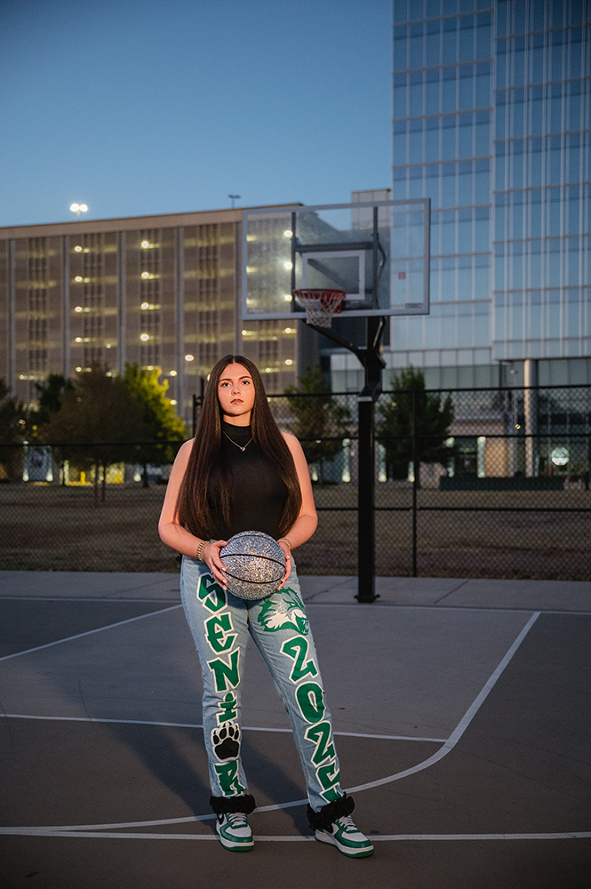 Peyton stands on an outdoor basketball court at dusk, holding a ball during her senior photo session. Her jeans say "Senior 2025." Buildings and hoop visible in the background.