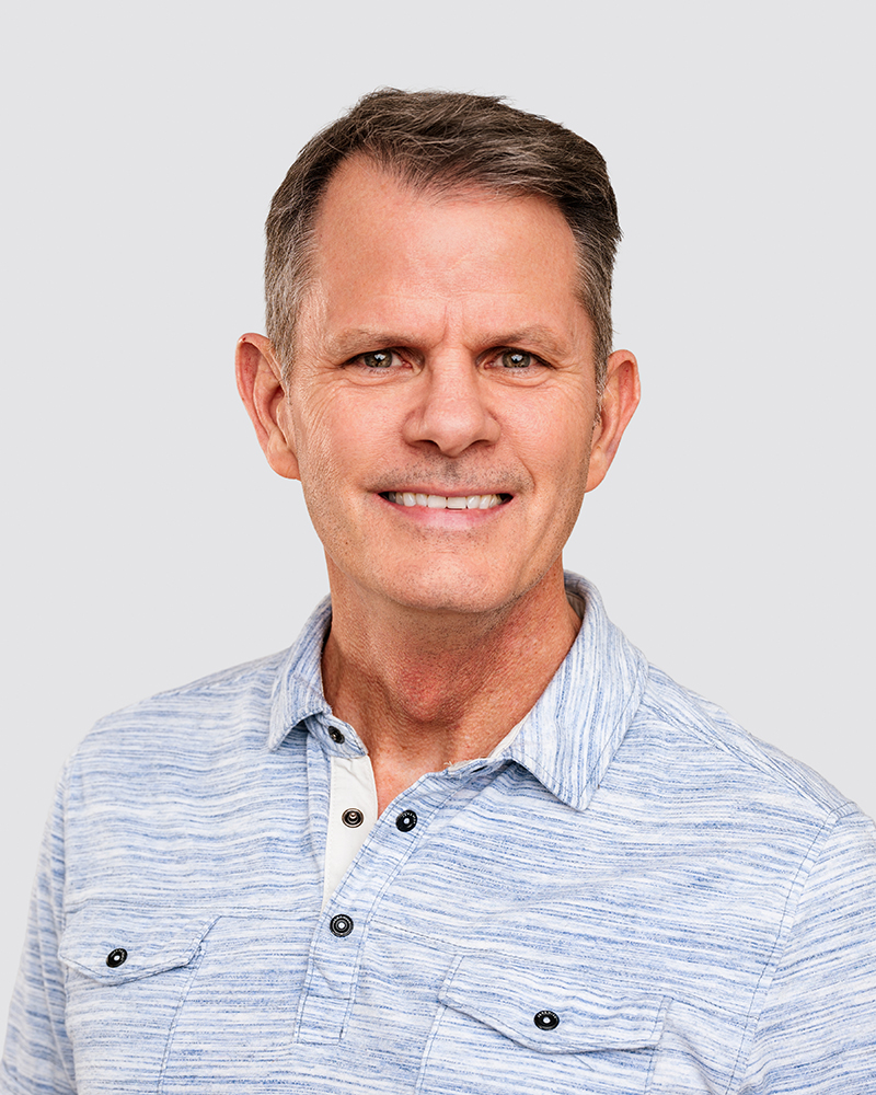 Headshot of John Wooley in a light blue, striped button-up shirt against a plain gray background. The image conveys a friendly, approachable tone.