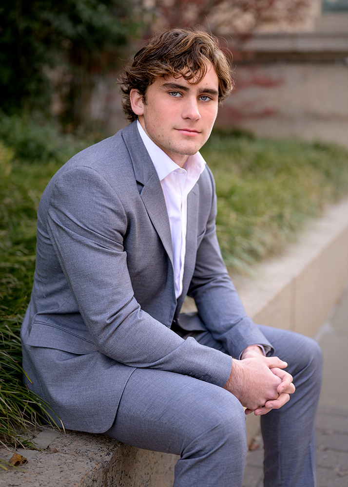 A high school senior in a gray suit sits on a stone ledge, hands clasped. He gazes forward with a confident expression, surrounded by greenery and a blurred background.