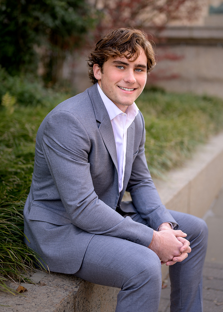 Smiling senior boy in a gray suit and white shirt sits on a stone ledge, surrounded by greenery. The scene conveys a professional and approachable tone.