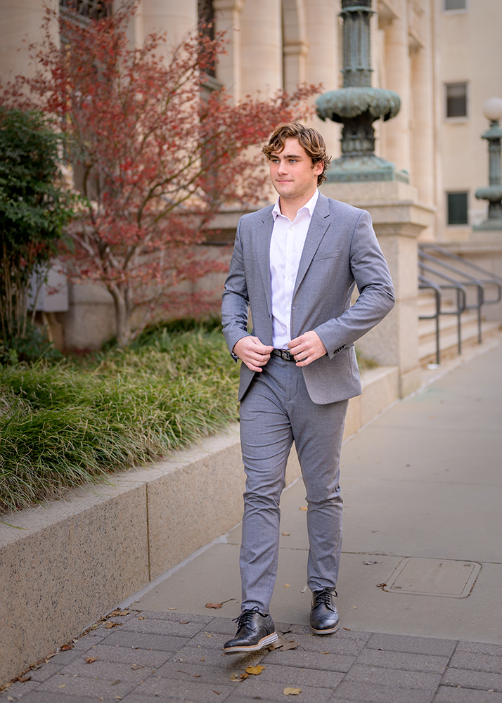 A high school senior in a gray suit and white shirt walks confidently on an OKC sidewalk, surrounded by autumnal trees and historic architecture, conveying a professional and calm demeanor.