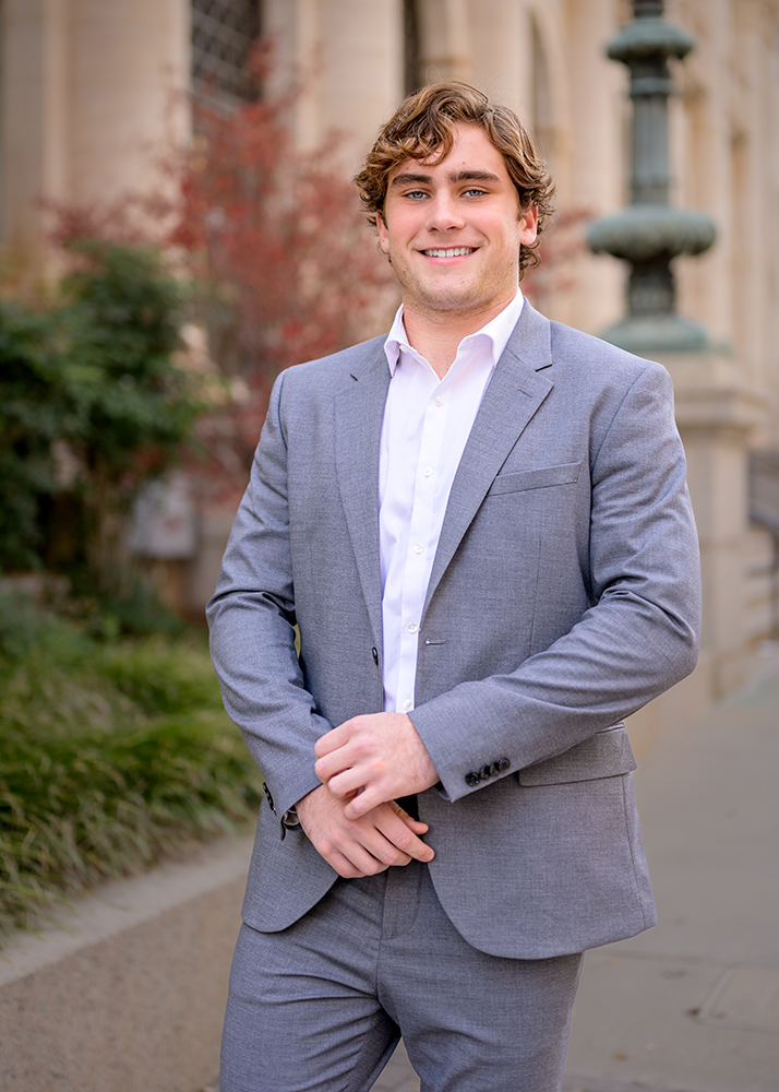 Senior guy in a gray suit and white shirt smiles confidently, standing outdoors with blurred greenery and a building in the background.
