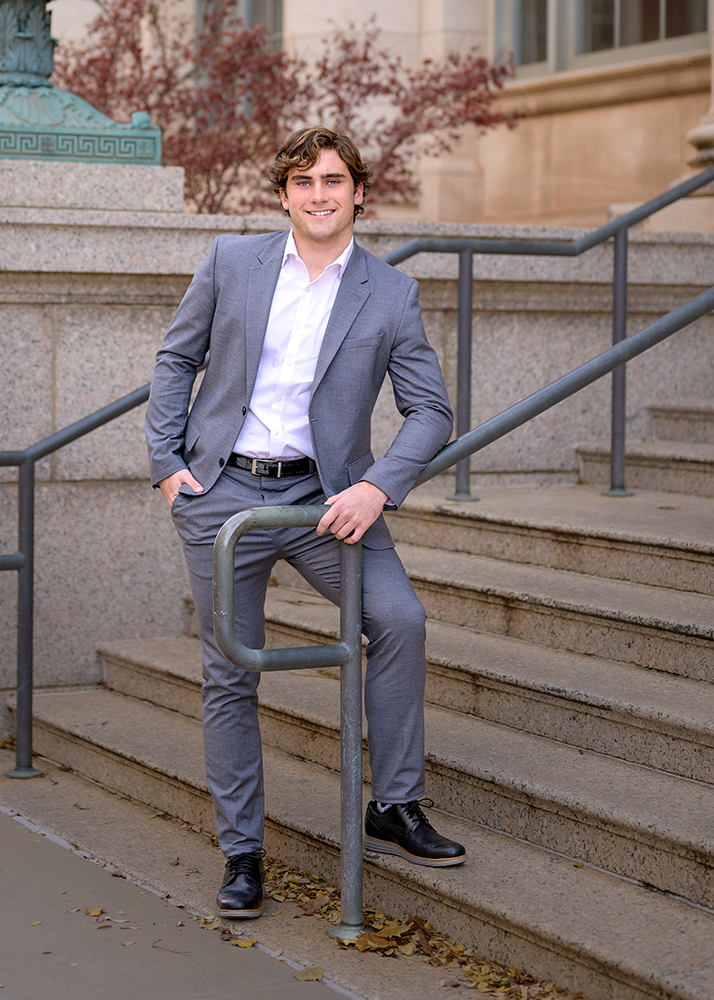 A smiling young senior guy in a gray suit and white shirt poses casually on stone steps. Autumn leaves dot the ground, and red foliage provides a warm backdrop.