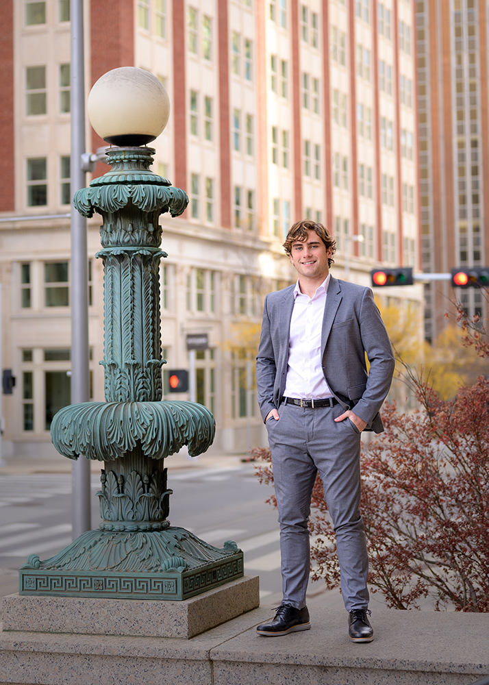 A high school senior in a gray suit and white shirt stands on a city street, smiling confidently. He is beside an ornate, turquoise lamp post, with a tall building in the background.