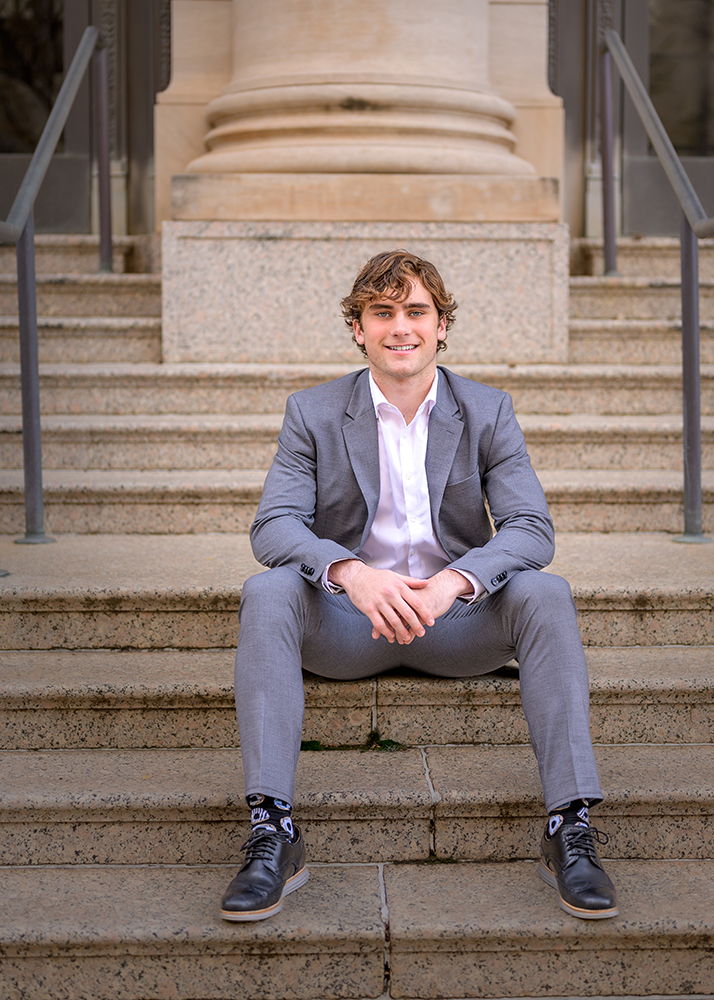 A senior boy in a gray suit and white shirt sits confidently on stone steps of a building, smiling. The background features stone columns and railings.