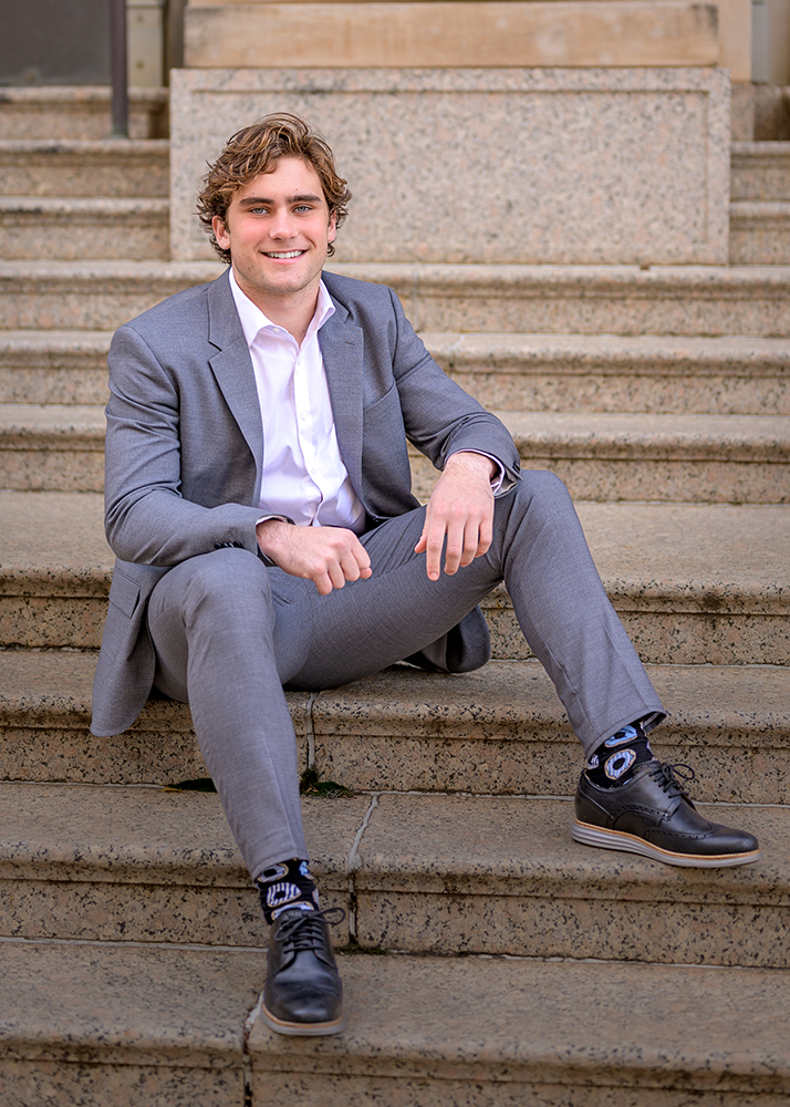 Smiling high school senior in a gray suit sitting casually on stone steps. He exudes confidence and warmth, with a friendly demeanor, wearing black shoes.