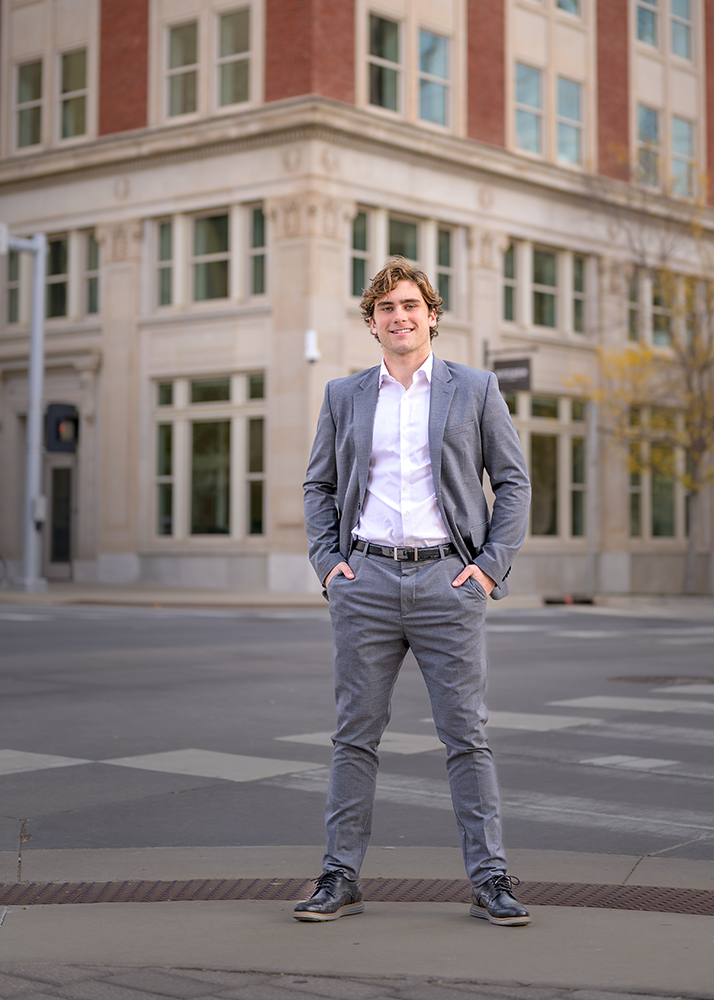 A high school senior in a gray suit and white shirt stands confidently on an urban street corner. The background shows a historic beige and red brick building.