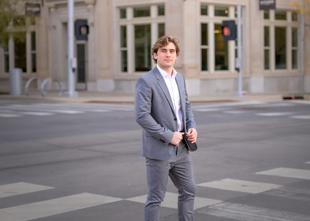 High school senior in a gray suit stands confidently at an urban crosswalk. A beige building with large windows is in the background. Daytime setting.