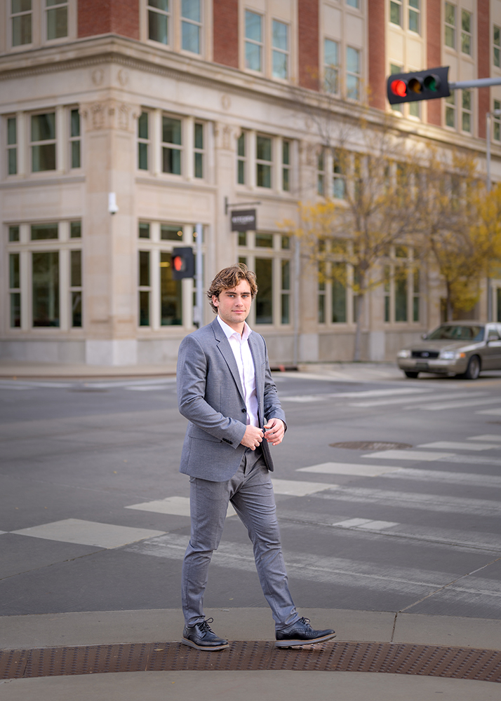 A high school senior in a gray suit crosses a city street. He's looking towards the camera with a neutral expression. The backdrop includes a brick building, trees with yellow leaves, and a car.