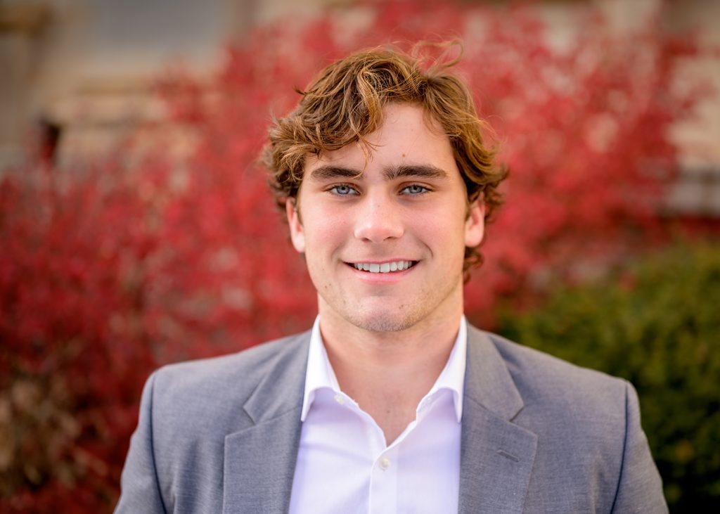 Young man with wavy hair smiles warmly. He's wearing a grey suit jacket over a white shirt. Red foliage blurs in the background, conveying a vibrant, cheerful tone.