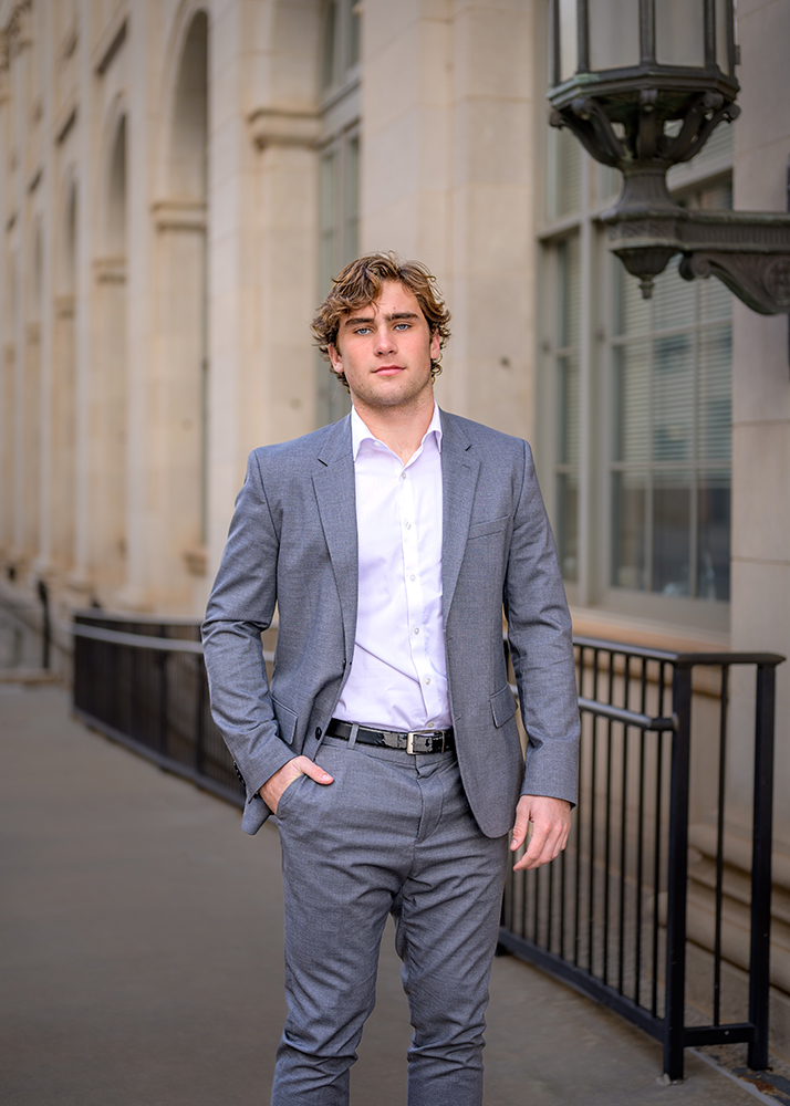 Senior guy in a gray suit stands confidently outside a stone building with arched windows, hand in pocket, exuding a professional and composed demeanor.