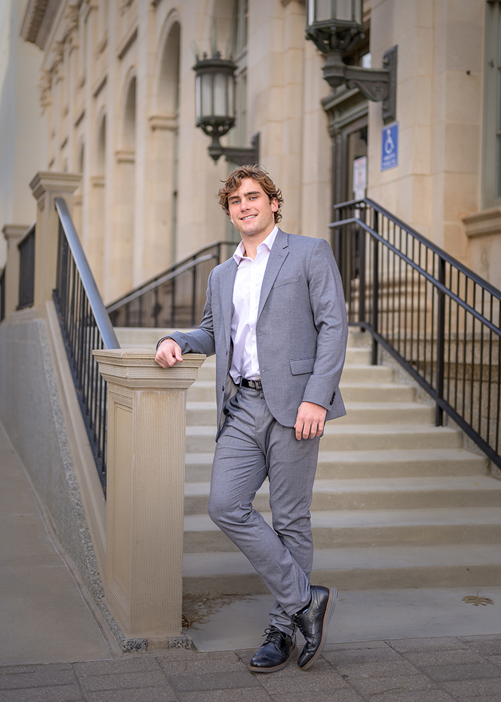 A high school senior in a gray suit leans against a railing in front of a historic building. He smiles confidently, conveying a professional and relaxed tone.