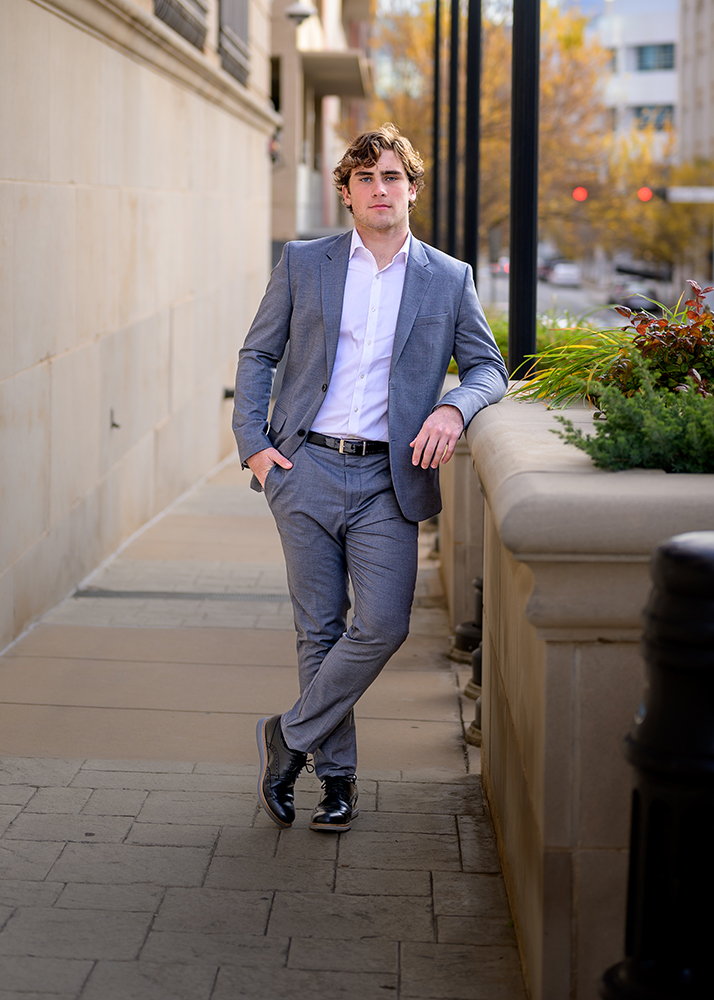Senior boy in a gray suit leans casually against a stone railing on a city sidewalk. He has a relaxed pose with autumn foliage in the background.
