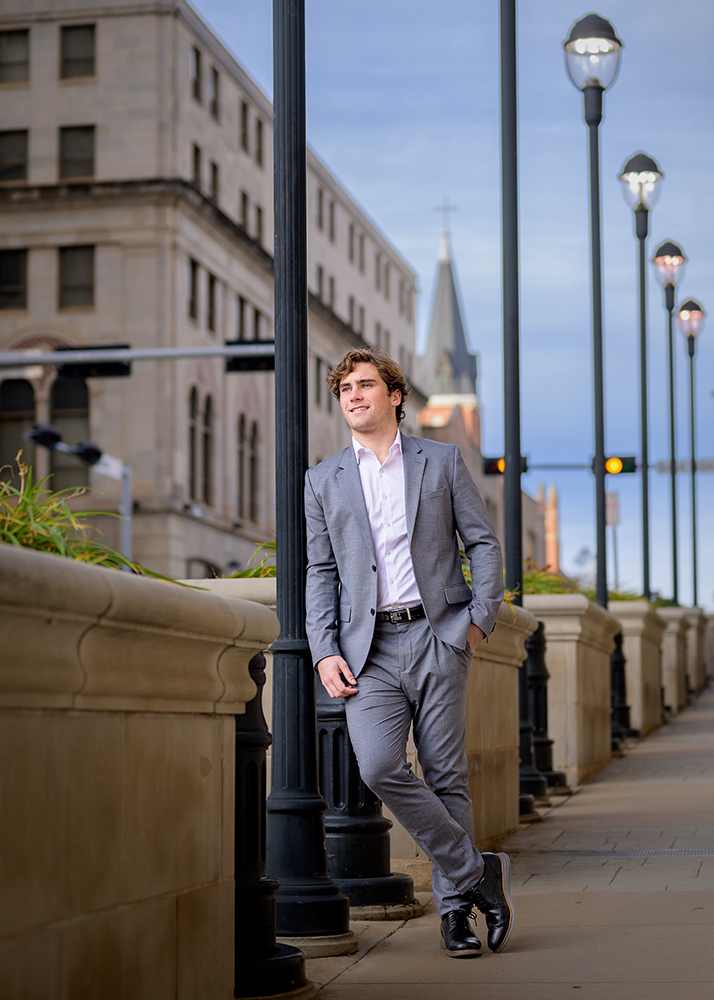 A high school senior in a gray suit leans against a lamppost, with historic buildings and a church steeple in the background. The tone is professional and confident.