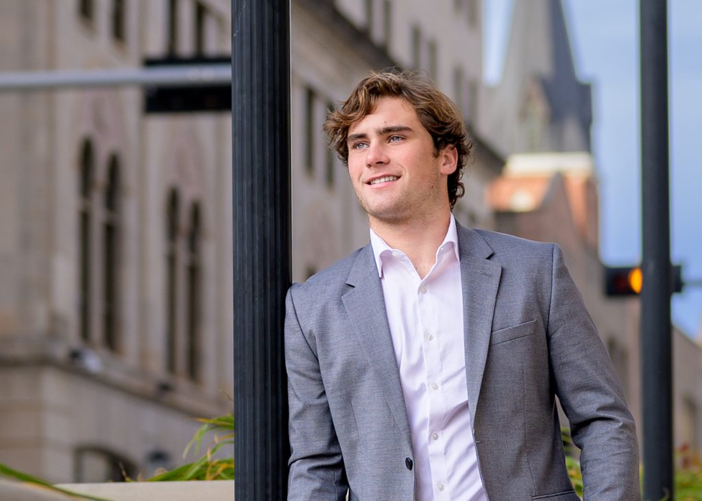 A high school senior in a gray suit and white shirt smiles, leaning against a lamppost in an urban setting. The mood is confident and relaxed.