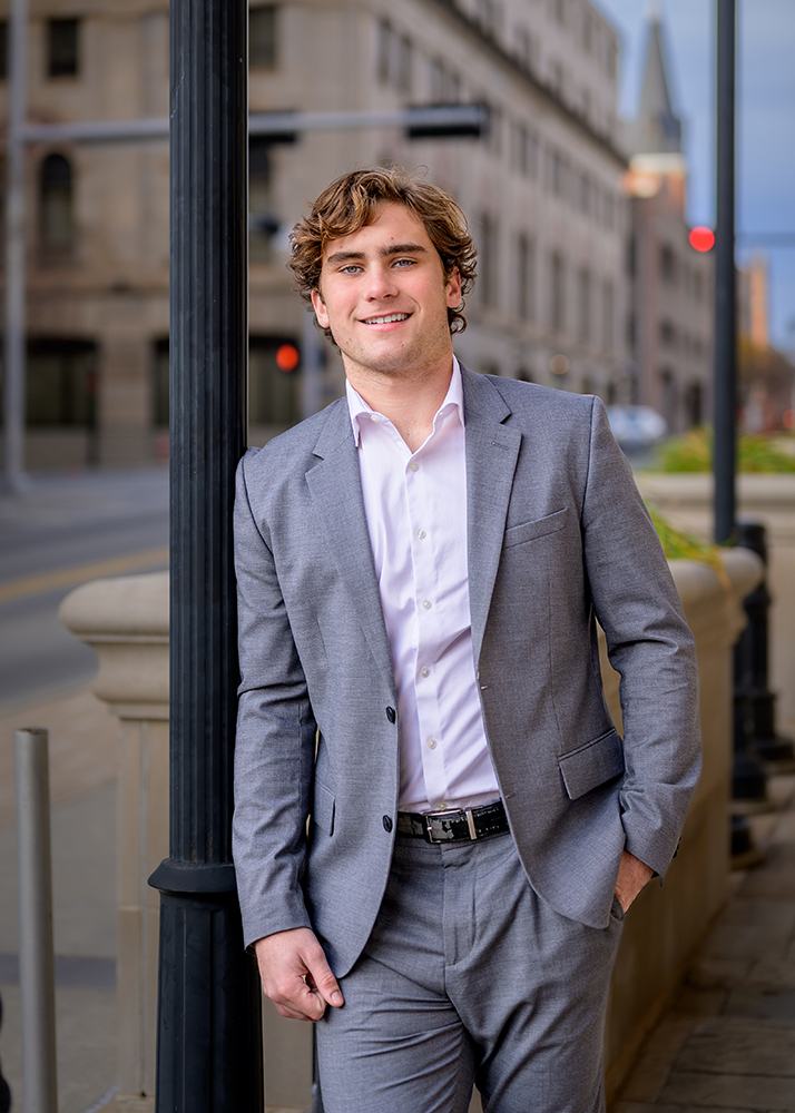 Senior guy in a gray suit and white shirt stands confidently against a black lamppost on a city street. Background shows a blurred urban scene.