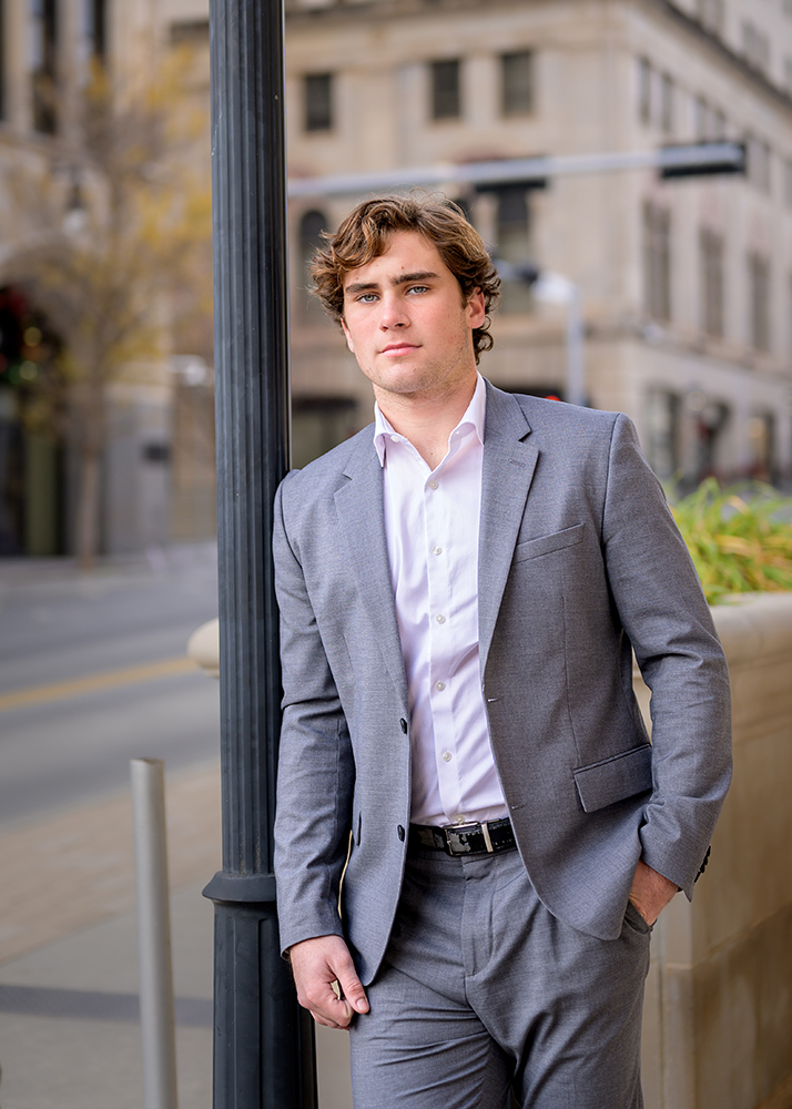Senior boy in a gray suit and white shirt stands confidently against a black lamppost on a city street. Background shows a blurred urban scene.