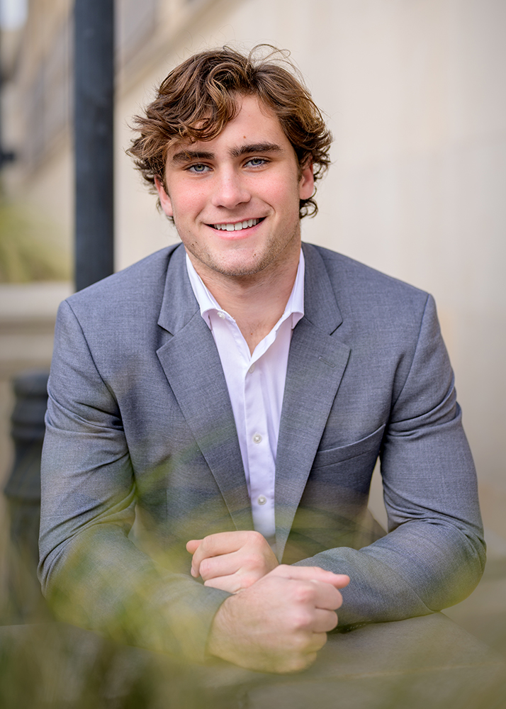 Jack wears a gray suit and white shirt smiles warmly, sitting in an outdoor setting. The background is softly blurred, creating a professional and approachable tone.