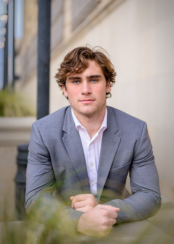 Young high school senior in a grey suit and white shirt, sitting outdoors against a blurred city backdrop. He appears confident and composed, looking directly ahead.