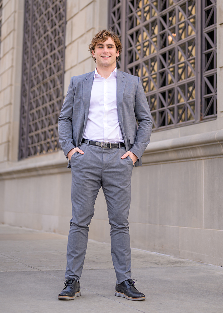 Young man in a grey suit and white shirt stands confidently against a patterned wall. He smiles warmly, hands in pockets, exuding a professional tone.