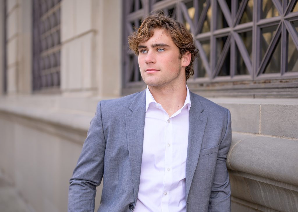 Senior boy in a gray blazer and white shirt leans against a stone building. He gazes confidently into the distance, exuding a professional demeanor.