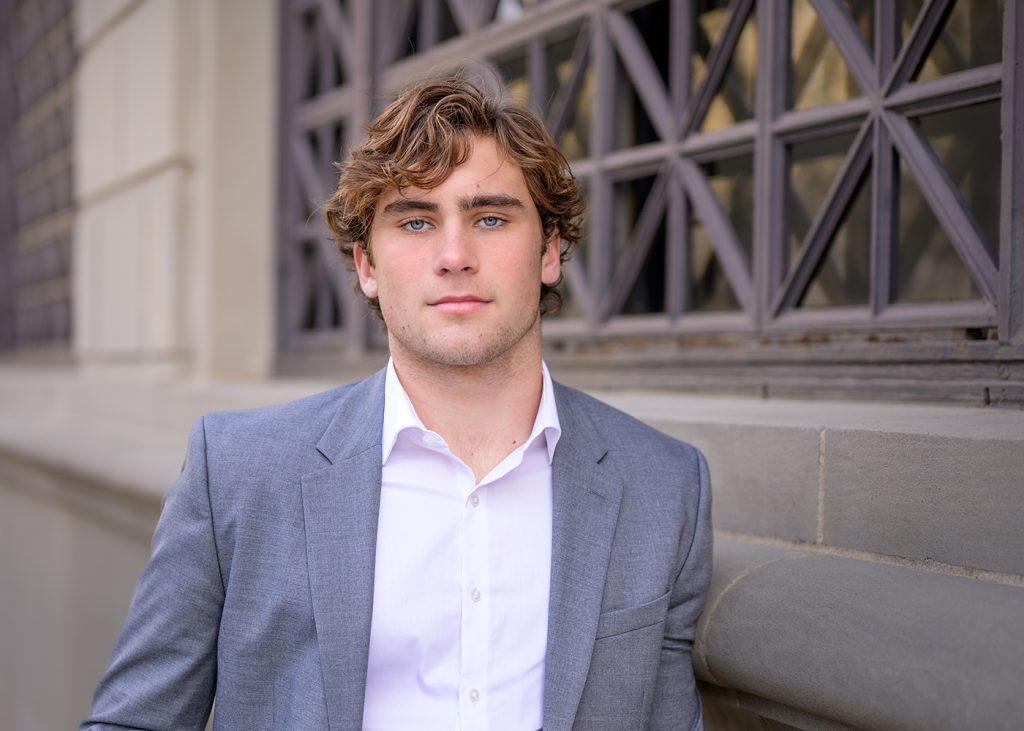 Young man with curly hair wearing a gray suit and white shirt, standing confidently in front of a building with decorative windows. Urban, professional tone.