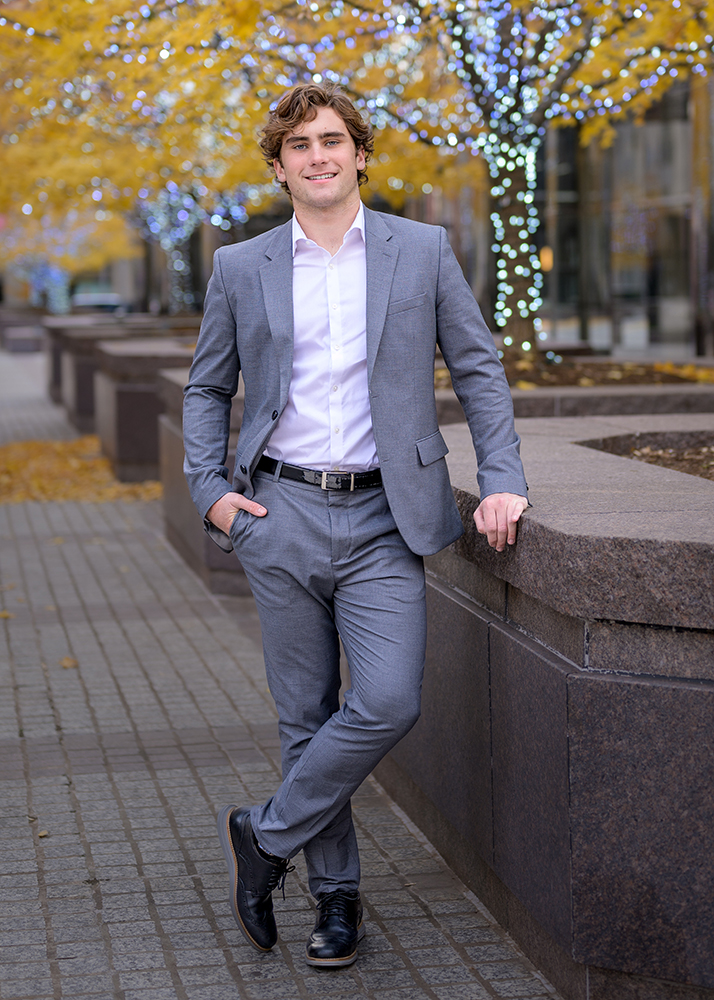 Young man in a gray suit and white shirt leans casually against a stone railing. Behind him, trees with yellow leaves and festive lights create a cheerful autumn scene.