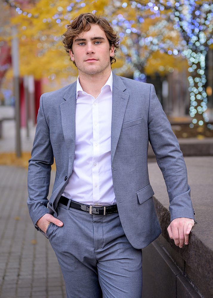 A high school senior in a light gray suit and white shirt stands confidently outdoors, with one hand in pocket. Background shows trees with autumn leaves and decorative lights.