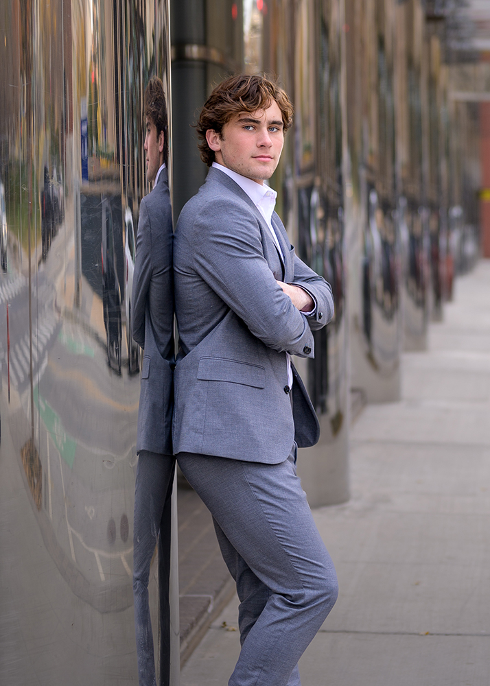 Senior guy in a gray suit leans against a reflective wall, arms crossed, exuding confidence. The urban setting features a blurred street view.
