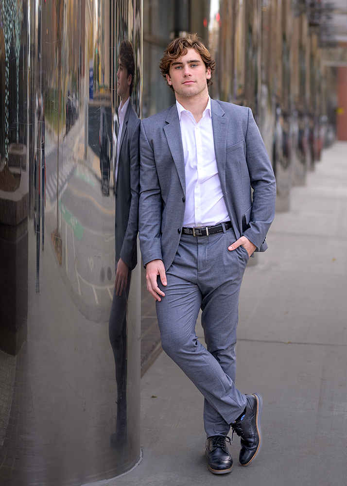 A high school senior in a gray suit and white shirt leans casually against a reflective building on a sidewalk. The street is visible, conveying a city vibe.