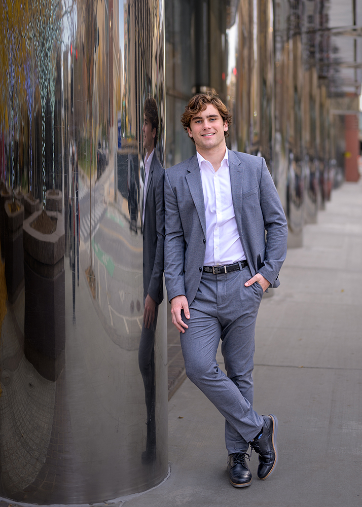 A high school senior boy in a gray suit and white shirt leans casually against a reflective building on a sidewalk. The street is visible, conveying a city vibe.