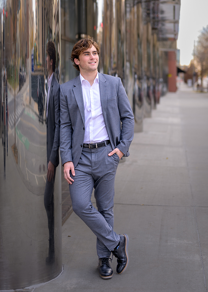 Smiling young man in a gray suit leans against a reflective building wall on an urban sidewalk during his senior photo session, exuding confidence and relaxation.