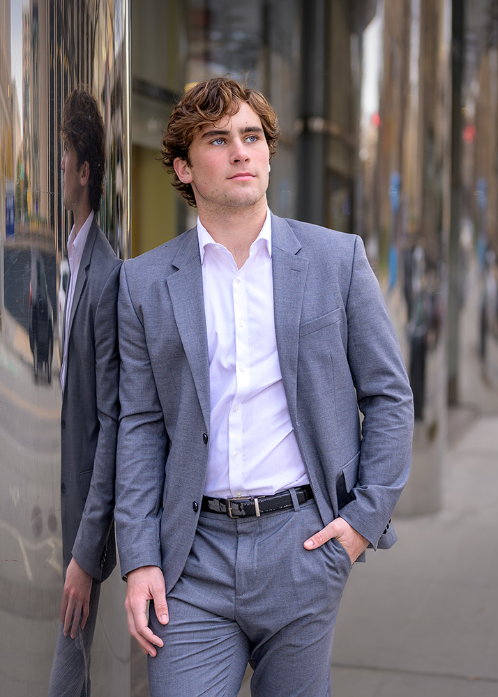 A young man in a gray suit and white shirt leans against a reflective building, looking contemplative. Urban background with soft focus.